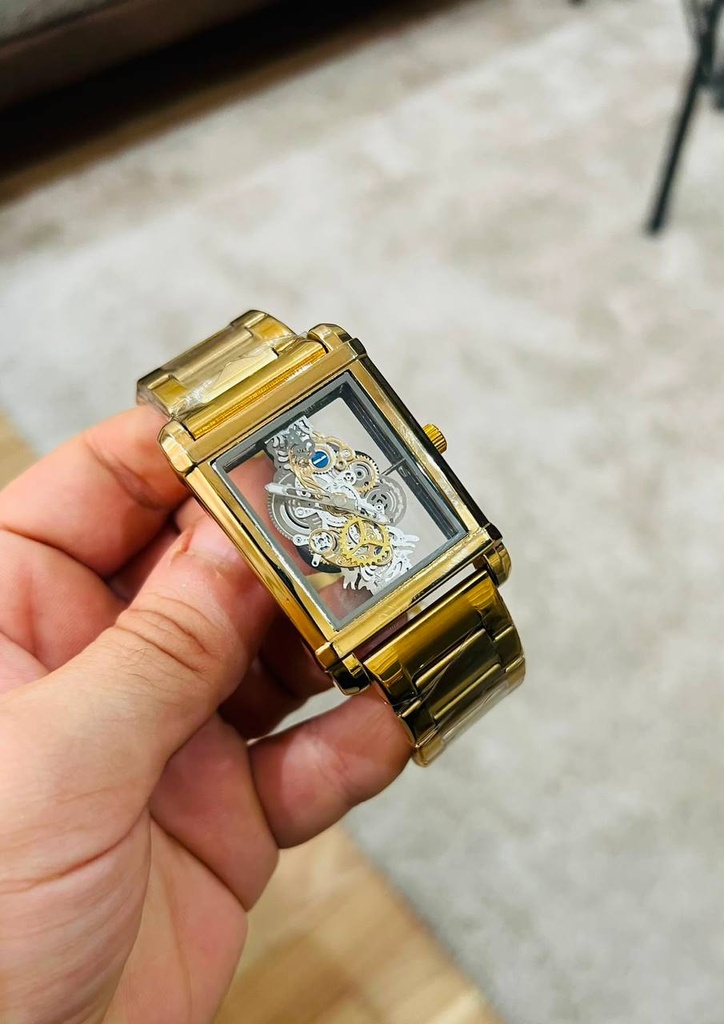 تظهر في الصورة ساعة يد رجالية فاخرة بتصميم مفرغ (Skeleton Watch) باللون الذهبي. تتميز الساعة بهيكل مستطيل زجاجي شفاف بالكامل يكشف عن تفاصيل التروس الميكانيكية الداخلية الدقيقة وميكانيزم الحركة بوضوح. تأتي الساعة مع سوار معدني (Steel) مطلي باللون الذهبي اللامع ومحمي بغلاف بلاستيكي شفاف يحافظ عليها جديدة)متوفر لون جولد واسود وسلفر: